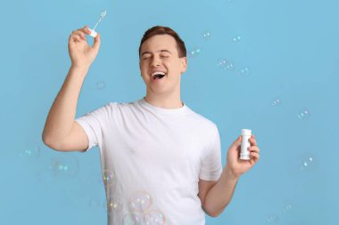 Genç mutlu yakışıklı adam mavi arka planda sabun köpüğüyle oynuyor.
