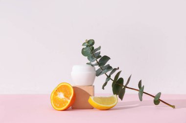 Turunçgil meyveleri, okaliptüs dalı, alçı standı ve pembe arka planda kozmetik ürün kavanozu içeren kompozisyon