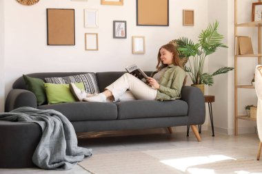 Oturma odasındaki kanepede dergi okuyan genç bir kadın.