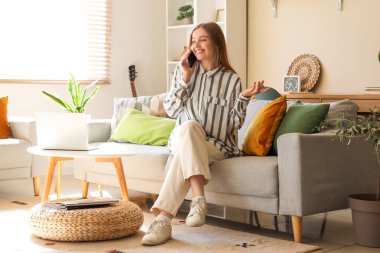 Kanepede oturan ve oturma odasında cep telefonuyla konuşan genç bir kadın.