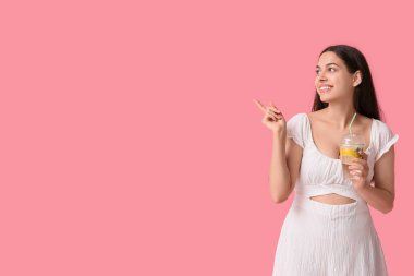 Bir fincan taze limonatayla pembe arka plandaki bir şeyi işaret eden güzel, genç ve mutlu bir kadın.