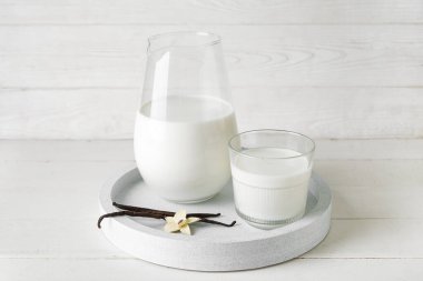 Bir bardak taze vanilya sütü, sopalı ve beyaz ahşap arka planda çiçek.