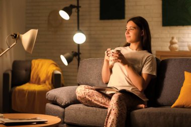 Pijamalı genç bir kadın dergi okuyor ve geceleri oturma odasında rahat bir kanepede çay içiyor.