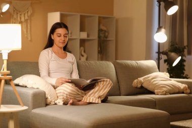 Pijamalı genç bir kadın geceleri oturma odasındaki yumuşak kanepede dergi okuyor.