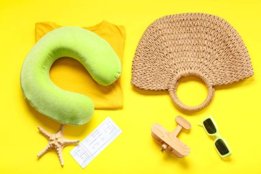 Seyahat yastığı, bilet, tişört ve sarı arka planda plaj aksesuarları.
