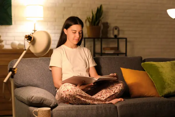 stock image Young woman in pajamas reading magazine on grey sofa in living room at night