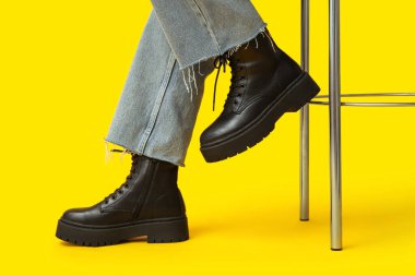 Sarı arka planda şık siyah deri çizmeli genç kadın bacakları.