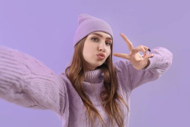 Örgü süveter ve şapka giymiş güzel genç bir kadın leylak arka planda selfie çekiyor.