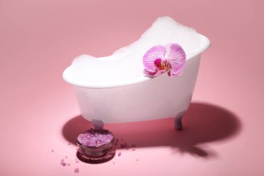 Köpüklü küçük küvet, pembe arka planda deniz tuzu ve orkide çiçeği.