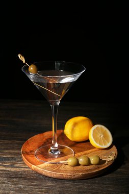 Lezzetli bir bardak martini, limon ve zeytinler ahşap masada, koyu arka planda.