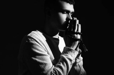 Tespihi olan genç adam karanlık arka planda dua ediyor.