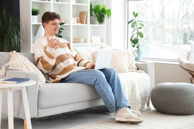 Bilgisayarlı erkek öğrenci evdeki kanepede ders çalışıyor.