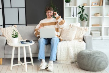 Kulaklıklı, dizüstü bilgisayarlı bir erkek öğrenci evdeki kanepede ders çalışıyor.