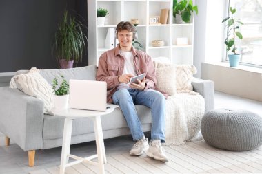 Kulaklıklı, tablet bilgisayarlı bir erkek öğrenci evdeki kanepede ders çalışıyor.