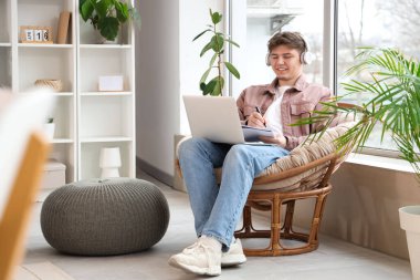 Kulaklıklı ve defterli bir erkek öğrenci evdeki koltukta ders çalışıyor.