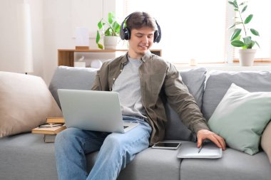 Kulaklıklı bir erkek öğrenci evdeki kanepede dizüstü bilgisayarla ders çalışıyor.