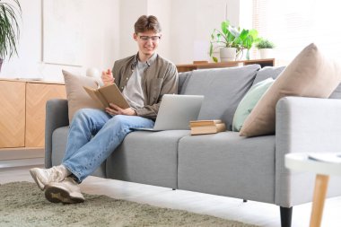 Kitabı olan bir erkek öğrenci evdeki kanepede ders çalışıyor.