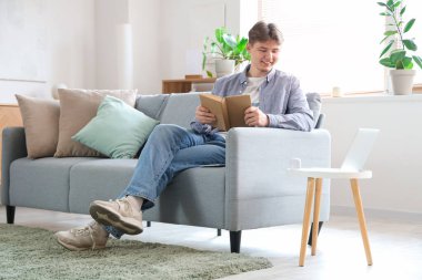 Kitabı olan bir erkek öğrenci evdeki kanepede ders çalışıyor.