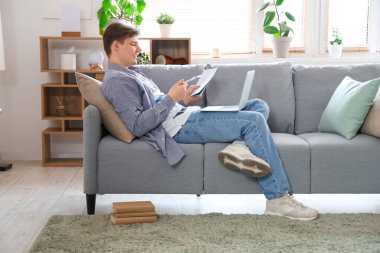 Not defteri ve dizüstü bilgisayarı olan erkek öğrenci evdeki kanepede ders çalışıyor.