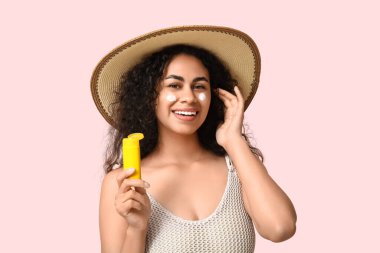 Pembe arka planda bir şişe güneş kremi olan güzel, mutlu, Afro-Amerikalı bir kadın.