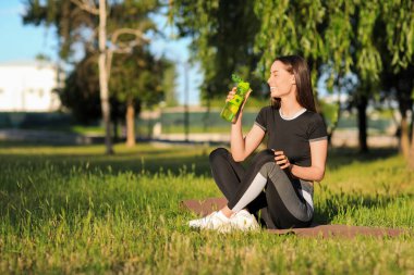 Parkta yoga minderinde oturan su şişeli genç bir kadın.