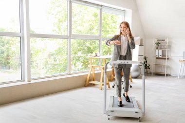 Ofisteki koşu bandında cep telefonlu genç bir iş kadını kol saatine bakıyor.