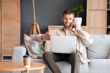 Bilgisayarlı yakışıklı adam evdeki koltukta cep telefonuyla konuşuyor.