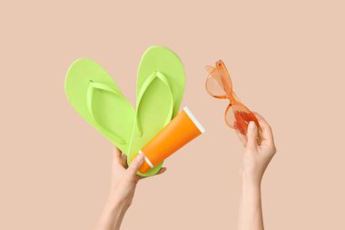 Güneş kremi, parmak arası terlik ve bej arka planda güneş gözlüğü olan kadın elleri.