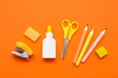 Bir şişe tutkal, makas ve turuncu arka planda keçeli kalemler. Üst görünüm