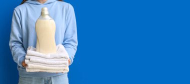 Mavi arka planda çamaşır deterjanı ve temiz havlu yığını tutan bir kadın.