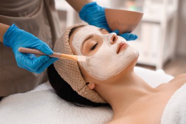 Güzellik salonunda genç bir kadının yüzüne maske takan kozmetikçi.