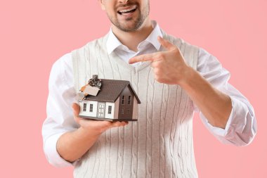 Erkek emlakçı ev modelini işaret ediyor. Pembe arka planda anahtarları var. Yakın plan.