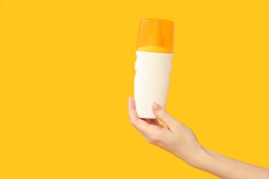 Sarı arka planda güneş kremi şişesi olan bir kadın eli.