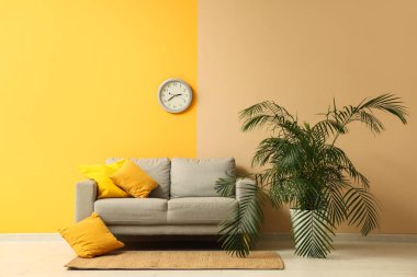 Şık bir oturma odası, rahat bir kanepesi, sarı duvarda çiçekleri ve saati var.