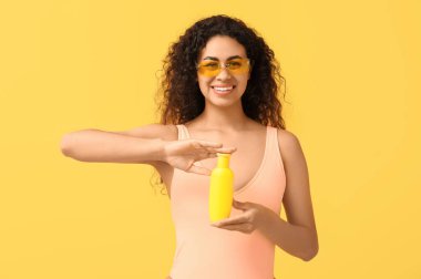 Sarı arka planda bir şişe güneş kremi olan güzel, mutlu, Afro-Amerikalı bir kadın.