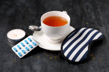 Uyku maskeli bir fincan çay, haplar ve siyah arka planda yanan mum.