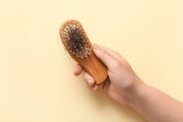 Рука держит расческу с куском волос на бежевом фоне. Проблема выпадения волос