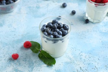 Taze yaban mersinli bir bardak lezzetli yoğurt ve açık mavi arka planda ahududu.