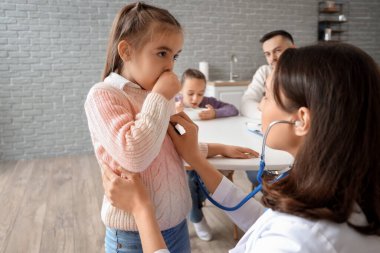 Female doctor listening lungs of ill little girl in kitchen