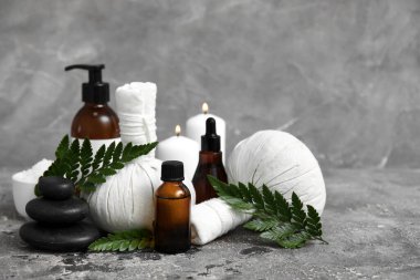 Bitkisel torbalar, deniz tuzu, spa taşları, eğrelti otu dalları ve gri arka planda yanan mumlarla farklı kozmetik ürünleri.