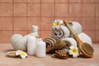 Masaj fırçaları, bitki torbaları, tesisat çiçekleri ve bej masa üzerinde yanan mumlar olan spa malzemeleri.