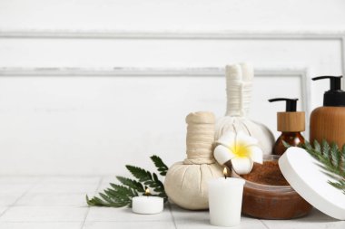 Farklı kozmetik ürünleri, bitkisel poşetler, eğrelti otu dalları, erik çiçeği ve beyaz fayans masasında yanan mumlar içeren spa malzemeleri.