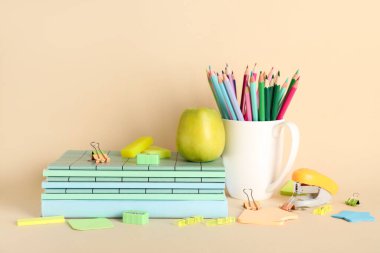 Bej arka planda farklı okul malzemeleriyle yeşil elma ve kalemler.