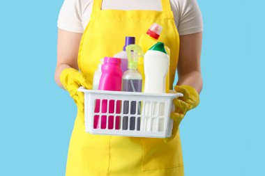 Mavi arka planda temizlik malzemesi sepeti olan kadın hademe, yakın plan.