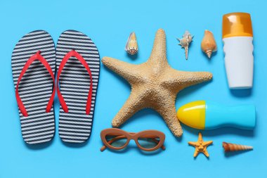 Şık çizgili terlikler, deniz kabukları, güneş gözlükleri, denizyıldızı ve mavi arka planda güneş kremi şişeleri.