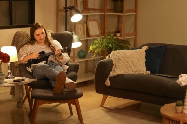 Genç ve güzel bir kadın akşam vakti koltukta oturup gitar çalıyor.