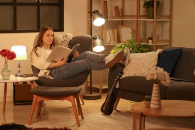 Genç ve güzel bir kadın akşam koltukta otururken dergi okuyor.