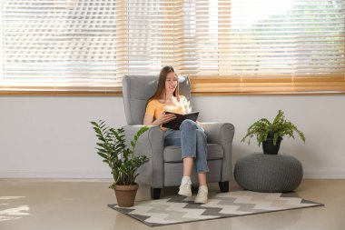 Odadaki rahat koltukta oturmuş kitap okuyan genç bir kadın.