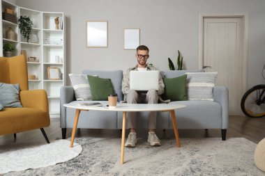 Gözlüklü genç adam evdeki kanepede dizüstü bilgisayar kullanıyor.