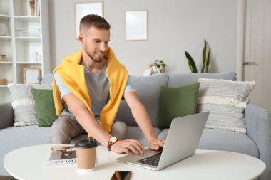 Genç sakallı adam evdeki masada dizüstü bilgisayar kullanıyor.